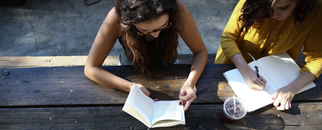 Zwei Junge Frauen sitzen an einem Holztisch im Freien. Die eine Frau liest, die andere schreibt etwas in ein Notizbuch.