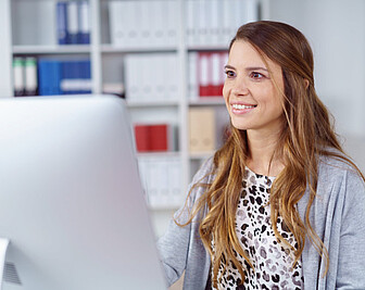 Foto: Eine Person sitzt an einem Schreibtisch und schaut auf einen Computerbildschirm. Im Hintergrund sind Regale mit Ordnern in verschiedenen Farben zu sehen. Die Person trägt ein gemustertes Oberteil und eine graue Strickjacke.