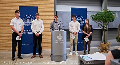 Foto: Fünf Personen stehen nebeneinander vor einem Rednerpult mit dem Logo der Universität Hohenheim. Zwei blaue Banner mit dem Universitätslogo sind im Hintergrund sichtbar. Eine Person hält ein Mikrofon, während die anderen Papiere in den Händen halten. Ein Baum steht rechts im Bild.