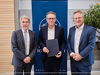 Foto: Drei Personen in Anzügen stehen vor einem blauen Banner mit einem Logo. Eine Person in der Mitte hält eine Urkunde. Der Text am unteren Rand erwähnt eine Preisverleihung 2025 an der Universität Hohenheim.