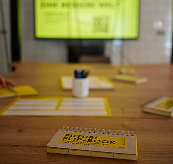Foto: Ein Tisch mit einem Notizbuch mit der Aufschrift 'FUTURE FLIP BOOK 5 Millionen Innovationen für eine bessere Welt'. Im Hintergrund ein unscharfer Bildschirm mit Text.