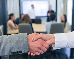 Foto: Eine Gruppe von Personen in formeller Kleidung steht in einem Raum und hält Smartphones. Sie scheinen in ein Gespräch vertieft zu sein. Im Hintergrund sind weitere Personen und unscharfe Bildschirme sichtbar. Die Umgebung wirkt wie ein Konferenz- oder Meetingraum.