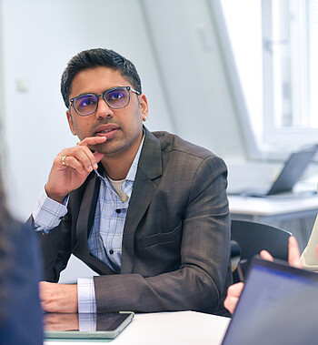 Foto: Eine Person in einem Anzug präsentiert vor einem Flipchart mit einem Balkendiagramm. Drei weitere Personen sitzen und hören zu. Das Diagramm zeigt Werte von 200 bis 300. Die Umgebung wirkt modern und hell.