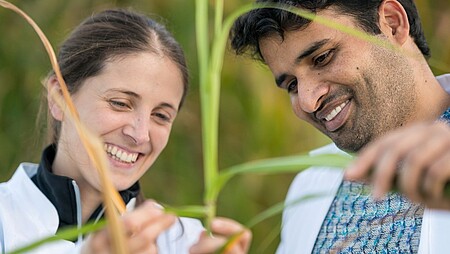 Zwei Personen, die in Wissenschaftskittel gekleidet sind, stehen in der freien Natur und untersuchen eine Pflanze.