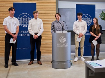 Foto: Fünf Personen stehen nebeneinander vor einem Rednerpult mit dem Logo der Universität Hohenheim. Zwei blaue Banner mit dem Universitätslogo sind im Hintergrund sichtbar. Eine Person hält ein Mikrofon, während die anderen Papiere in den Händen halten. Ein Baum steht rechts im Bild.