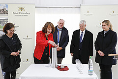 Coins from the donors: State Secretary Gisela Splett (Ministry of Finance BaWü) deposits euro coins into a time capsule for the foundation stone of the future animal science research center HoLMiR. The capsule also included construction plans and a current daily newspaper, deposited by Ministerial Director Dr. Hans J. Reiter (Ministry of Science BaWü, to the right of Splett), the Hohenheim guidelines for animal experiments, deposited by University President Prof. Dr. Stephan Dabbert (to the right of Reiter), and a Bezoar stone, deposited by agricultural scientist Prof. Dr. Jana Seifert (to the right of Dabbert). Bezoars are balls of animal hair and other foreign bodies that accumulate in animals’ stomachs. Pictured on the left: Carmen Zinnecker-Busch, from the Stuttgart and Hohenheim University Building Office. Image: University of Hohenheim/Beqiri Coins from the donors: State Secretary Gisela Splett (Ministry of Finance BaWü) deposits euro coins into a time capsule for the foundation stone of the future animal science research center HoLMiR. The capsule also included construction plans and a current daily newspaper, deposited by Ministerial Director Dr. Hans J. Reiter (Ministry of Science BaWü, to the right of Splett), the Hohenheim guidelines for animal experiments, deposited by University President Prof. Dr. Stephan Dabbert (to the right of Reiter), and a Bezoar stone, deposited by agricultural scientist Prof. Dr. Jana Seifert (to the right of Dabbert). Bezoars are balls of animal hair and other foreign bodies that accumulate in animals’ stomachs. Pictured on the left: Carmen Zinnecker-Busch, from the Stuttgart and Hohenheim University Building Office. Image: University of Hohenheim/Beqiri